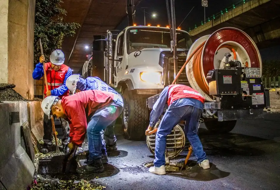 Street workers cleaning drainage at night with machinery under a bridge.